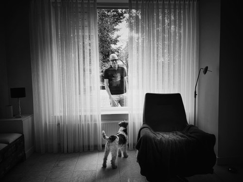 Jacques van Gerven kijkt van buiten door het raam een woonkamer in waarin een hond staat.